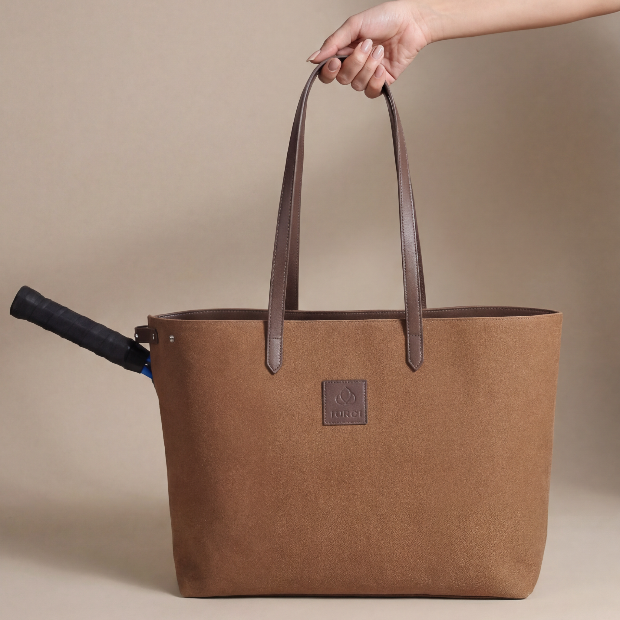 A hand holds a brown canvas and leather tennis tote with dark handles. A tennis racquet handle sticks out from the double-racket compartment on one side of the bag. The background is light brown.