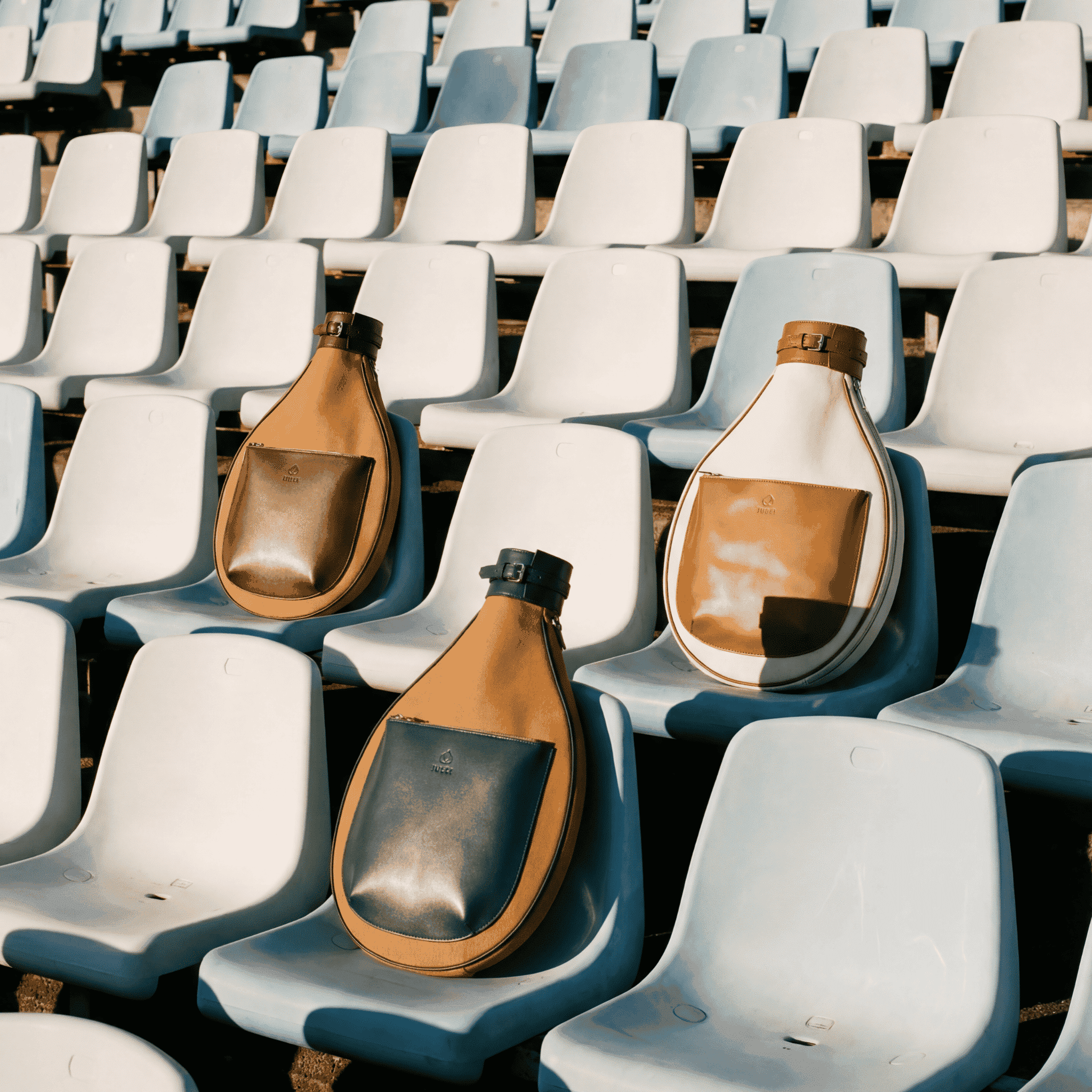 Three large, leather tennis bags in different colors rest on light blue plastic stadium seats in rows, with sunlight casting shadows across the otherwise empty seats.