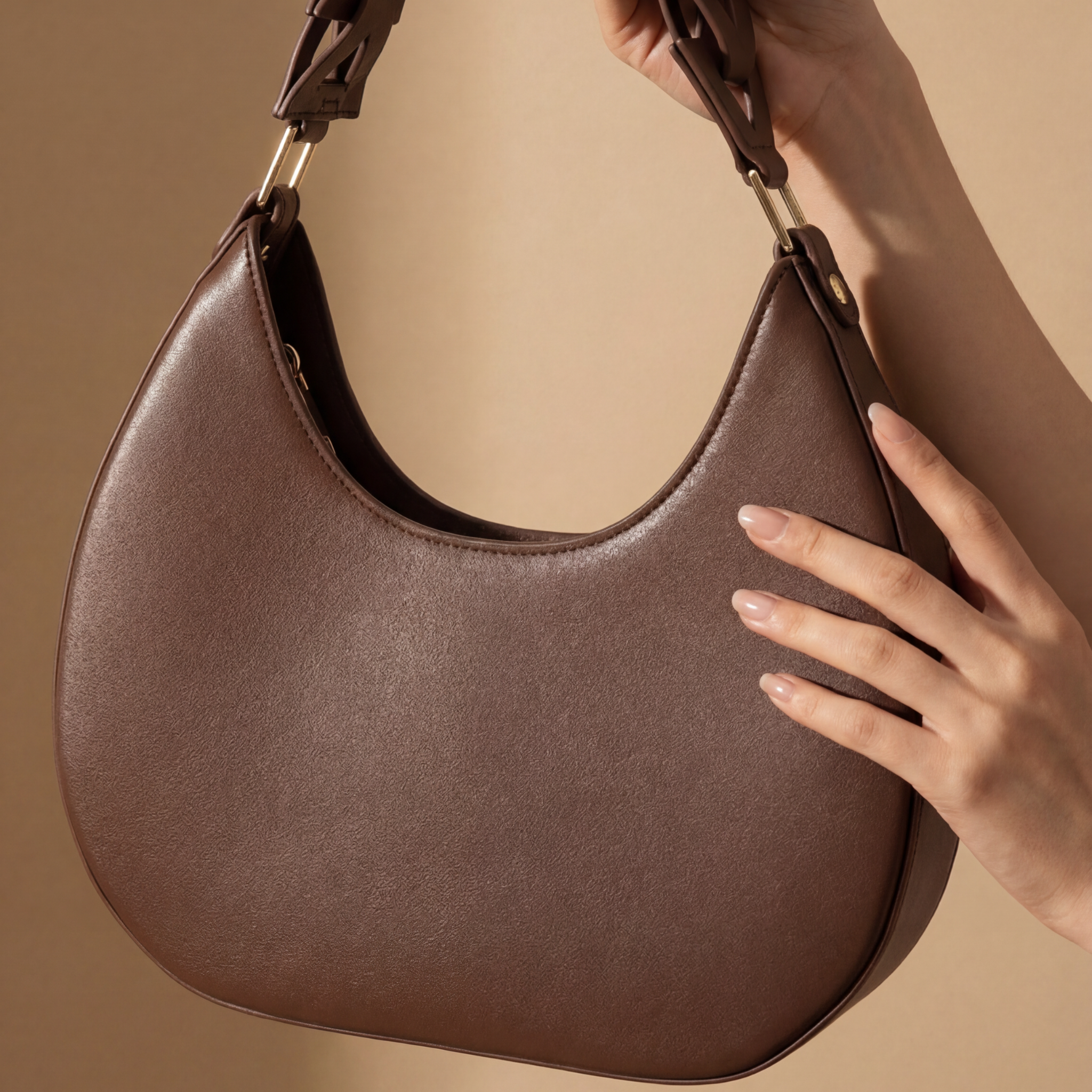 A person with manicured nails holds a brown D-Letter Bag with gold hardware against a beige background.