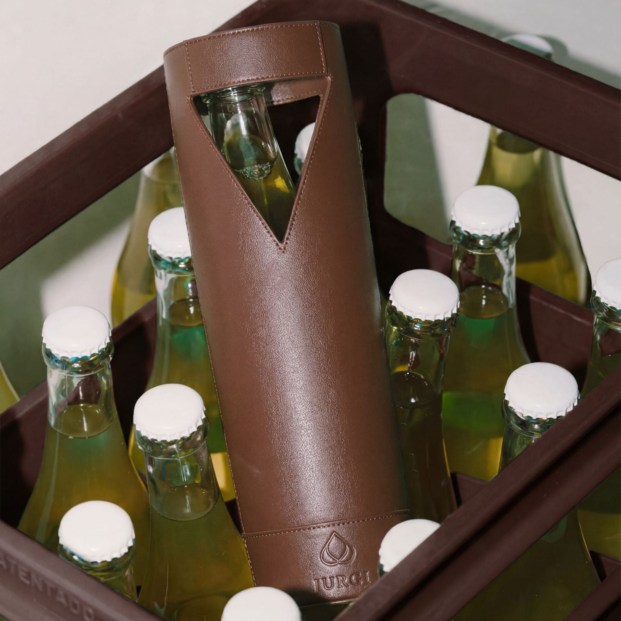 A brown leather wine bag with a triangular cutout displays a glass bottle inside, standing upright in a crate filled with similar capped glass bottles containing a yellow-green liquid.