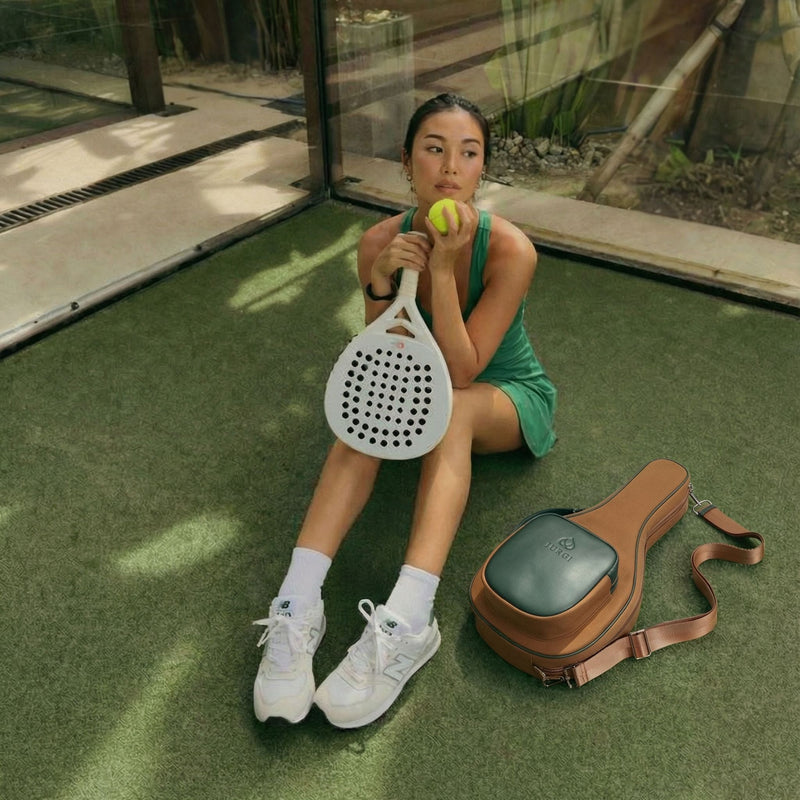 Woman sitting on a green surface holding a paddle and tennis ball, with a brown bag beside her.