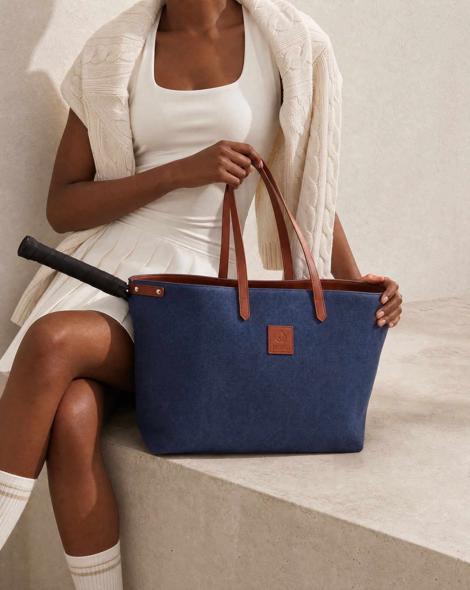 Navy canvas tote bag with brown leather handles and logo patch, shown with tennis racket, minimalist and elegant design.