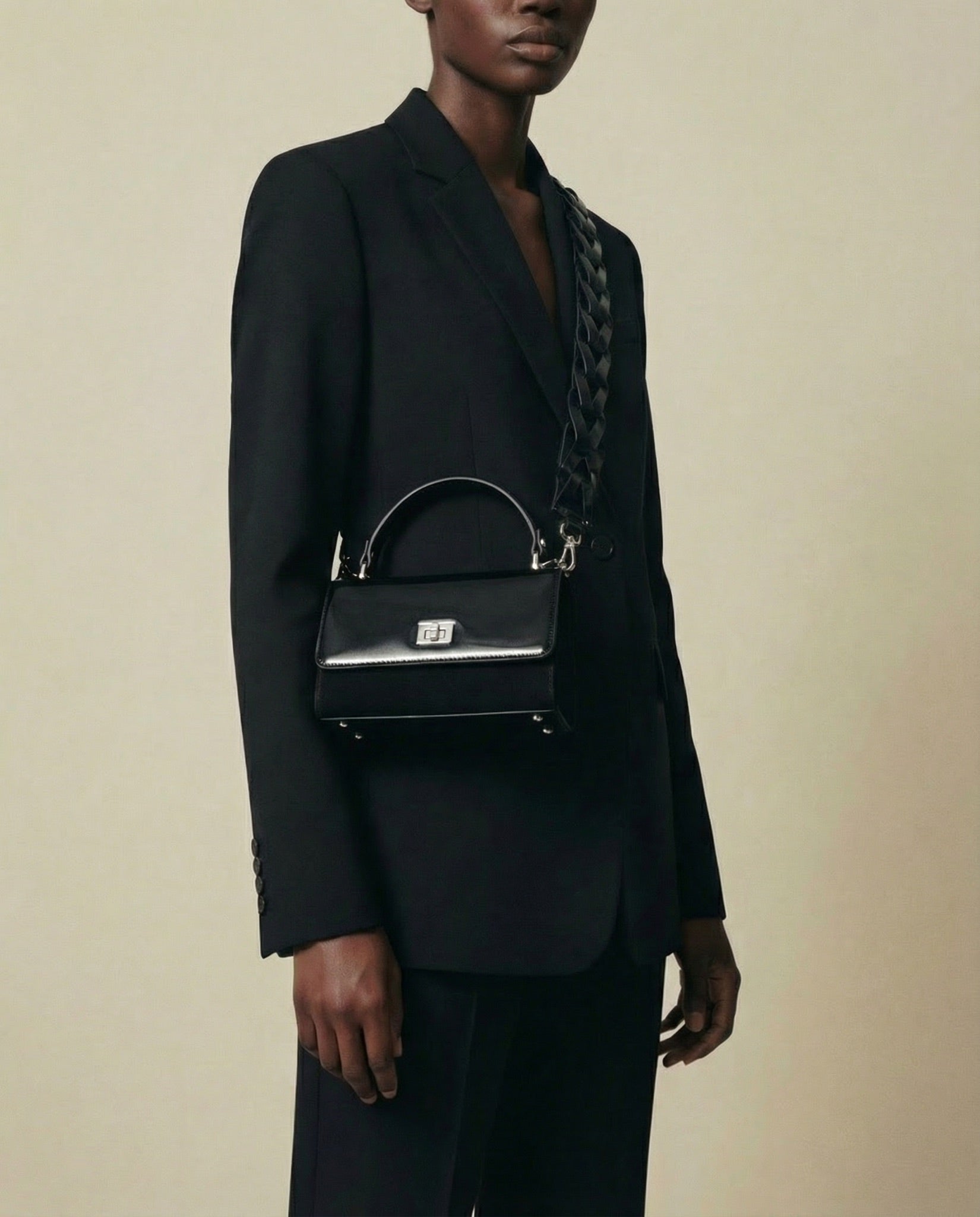 A person wearing a black suit holds a small black handbag with a braided shoulder strap and metallic clasp, standing against a plain beige background.
