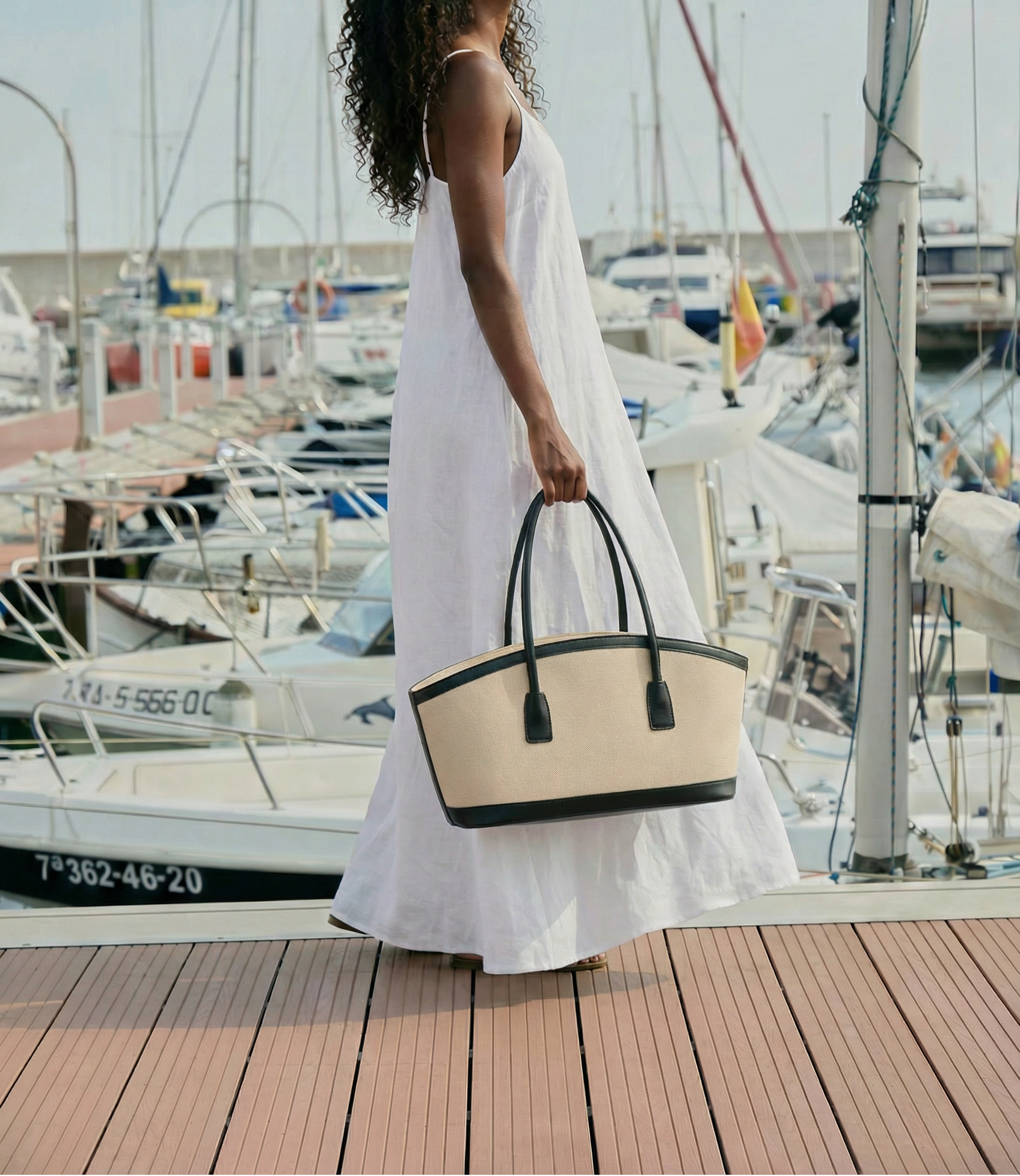 Woman in a white dress holding a beige and black handbag on a dock with boats in the background