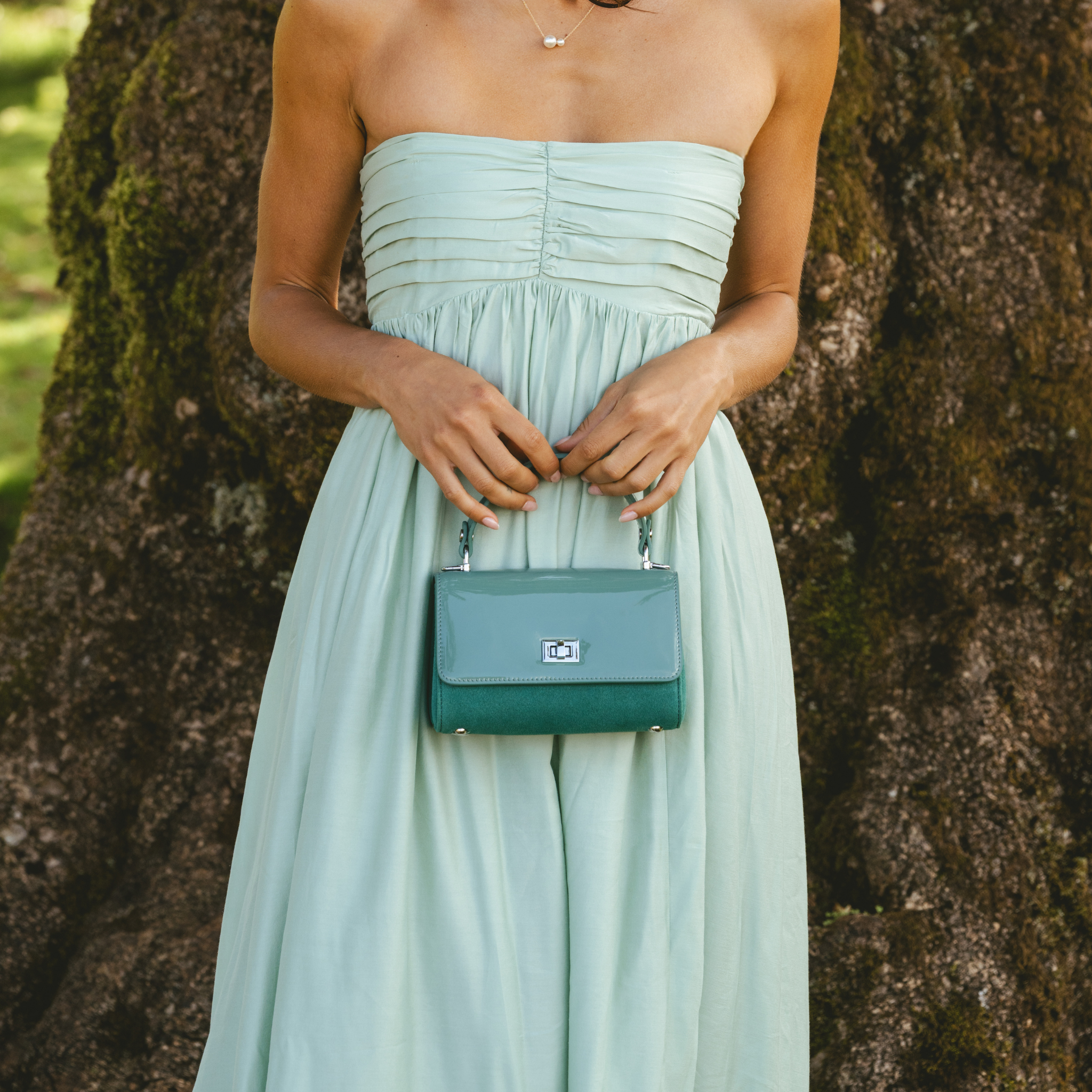 Woman in a light green strapless dress holding a teal handbag against a tree background