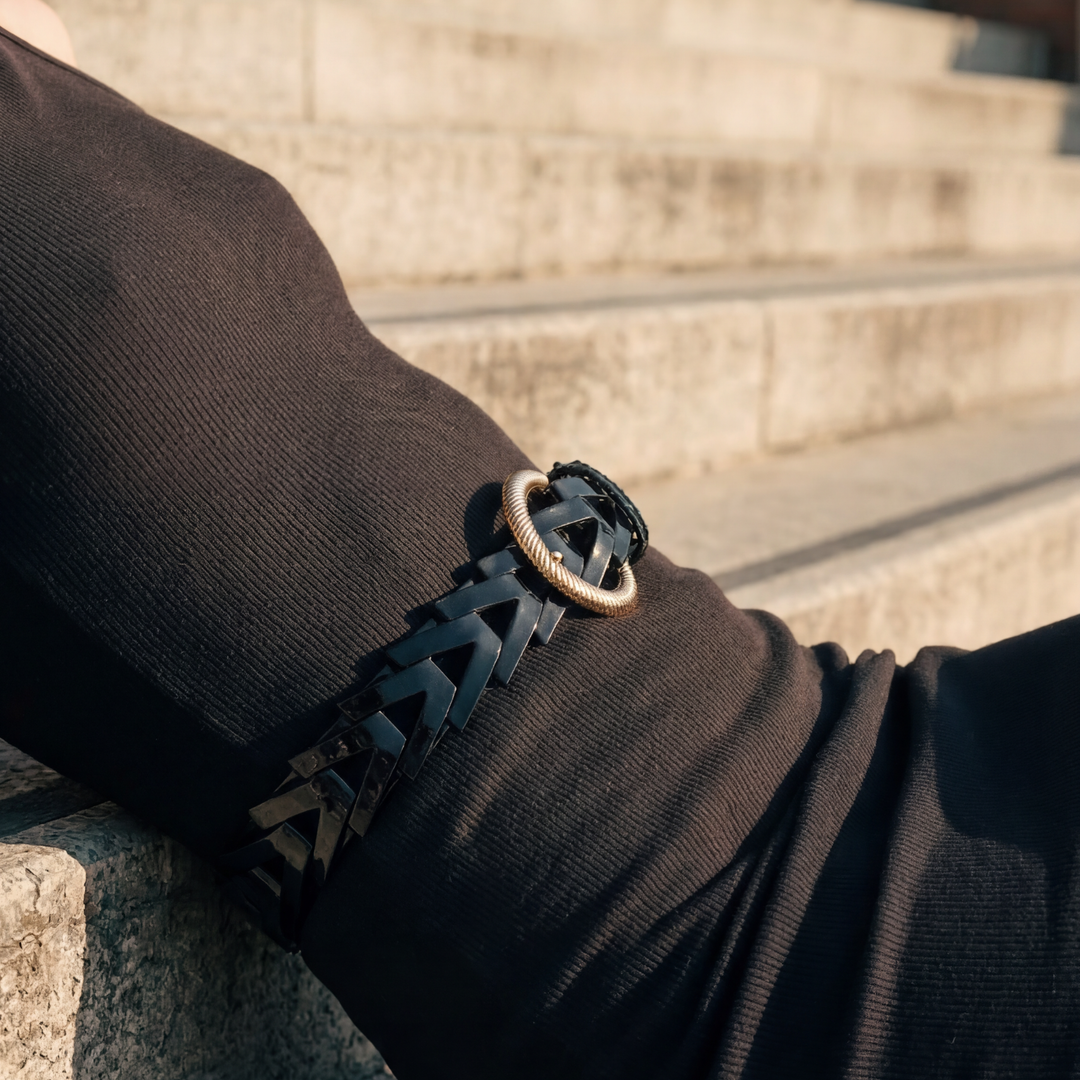 Close-up of a person wearing a black leather bracelet with a gold clasp on a blurred background.