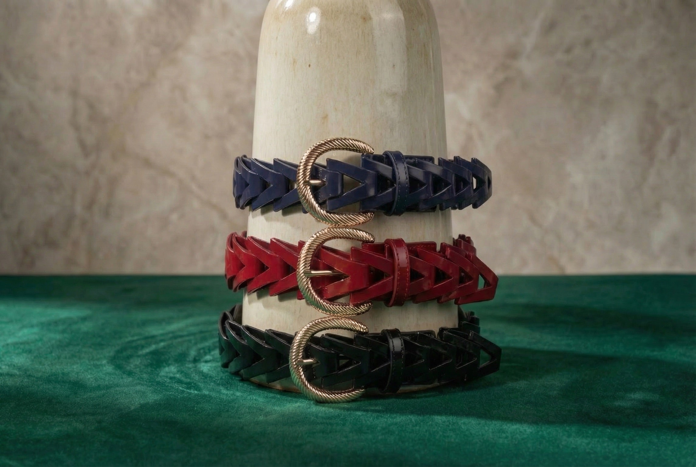Three braided leather belts, two navy and one red, each with a gold textured buckle, are wrapped around an off-white ceramic vase, set on a green velvet surface with a neutral backdrop.