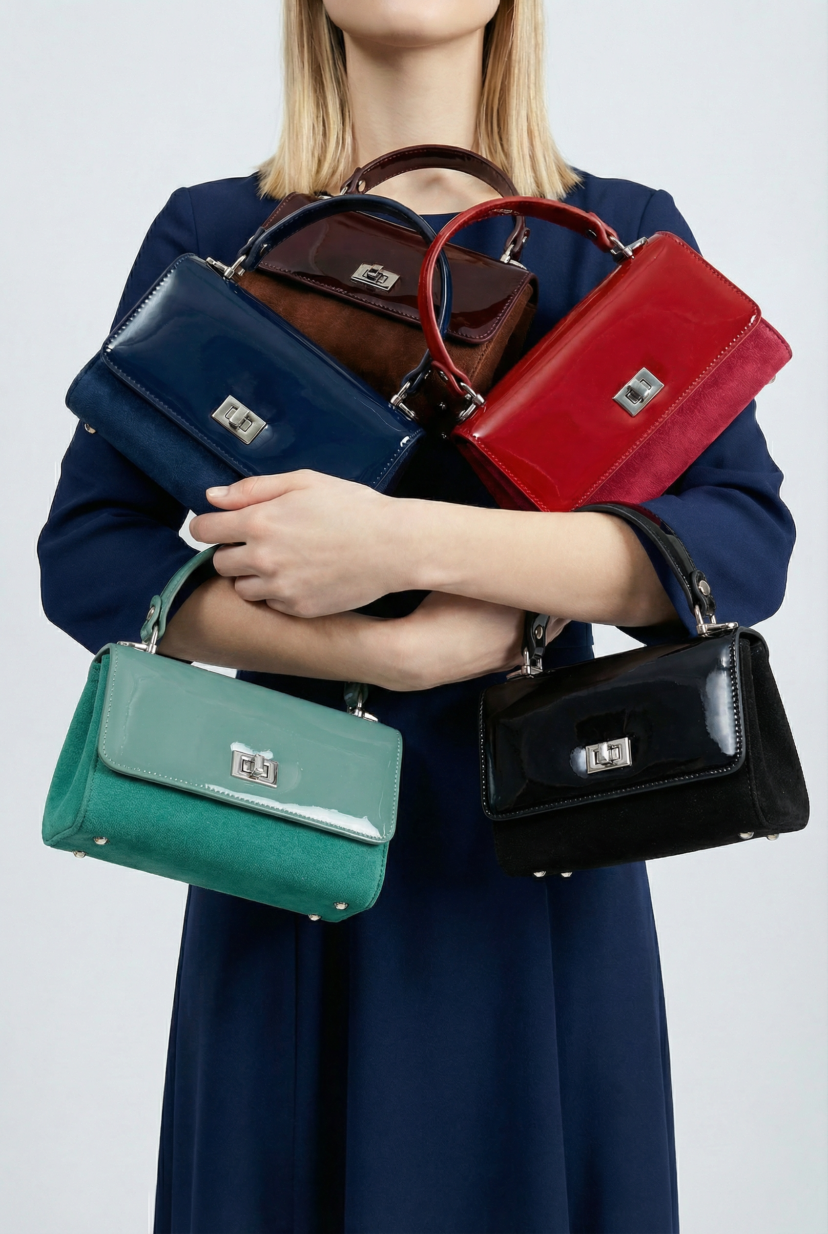 A woman in a navy blue dress holds five handbags in different colors—navy blue, brown, red, green, and black—stacked in her arms, with her face partially out of frame, against a plain background.